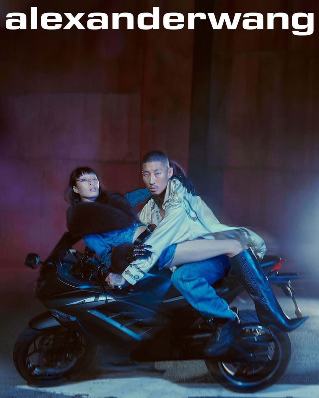 Two fashion models pose on a motorcycle at night under the Alexander Wang logo in an editorial image with cool color grading.