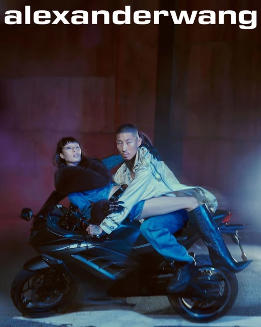 Two fashion models pose on a motorcycle at night under the Alexander Wang logo in an editorial image with cool color grading.