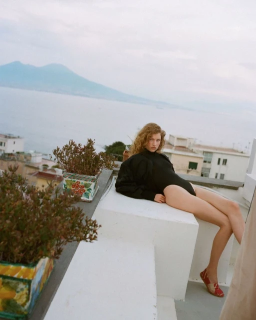Woman in black bodysuit poses on white rooftop with potted plants, overlooking the ocean and Mount Vesuvius on a cloudy day.