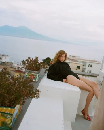Woman in black bodysuit poses on white rooftop with potted plants, overlooking the ocean and Mount Vesuvius on a cloudy day.