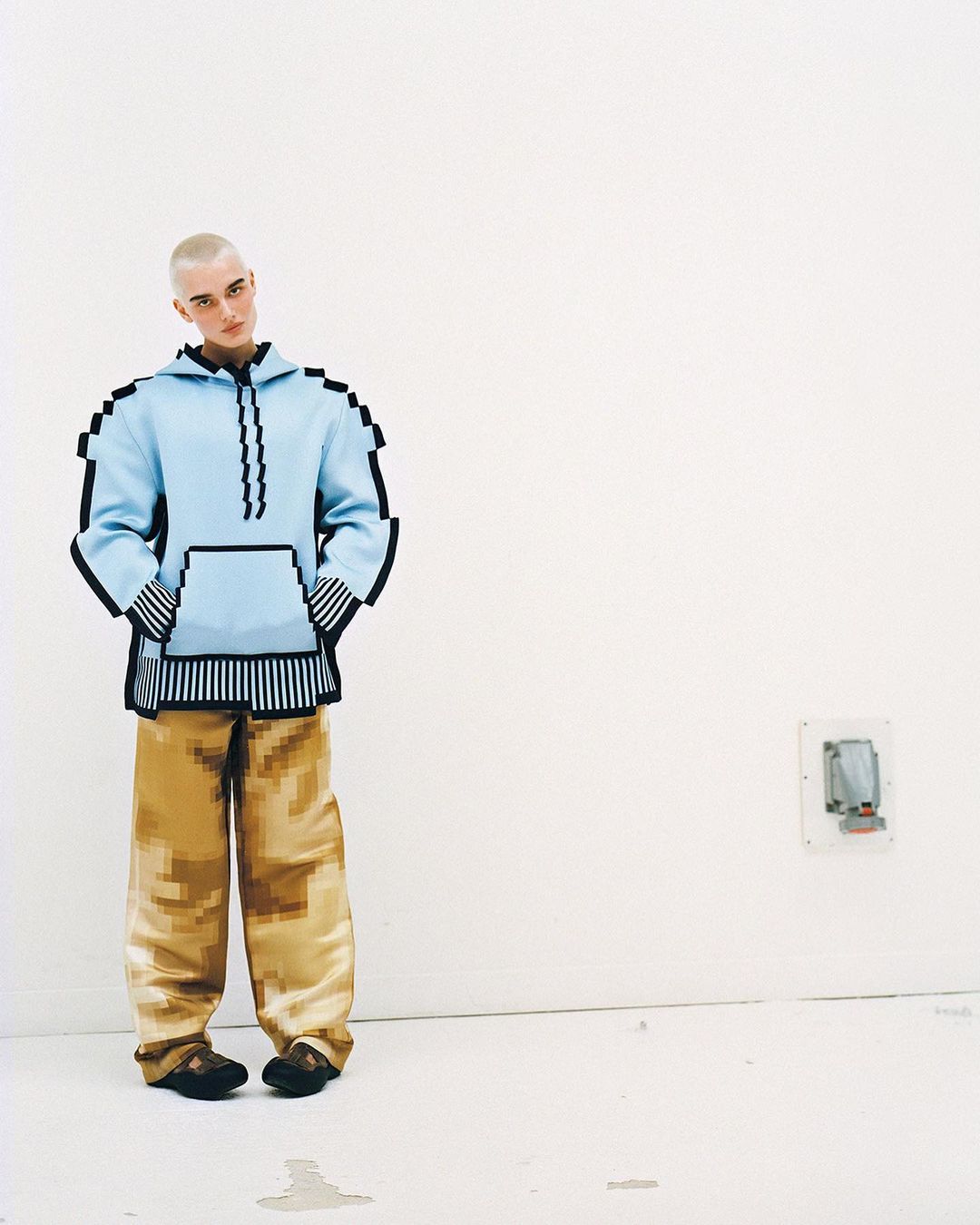 Person in pixelated fashion: light-blue hoodie with black trim and beige pants, standing against a white wall in a minimalist room.