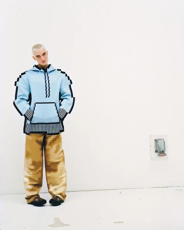 Person in pixelated fashion: light-blue hoodie with black trim and beige pants, standing against a white wall in a minimalist room.