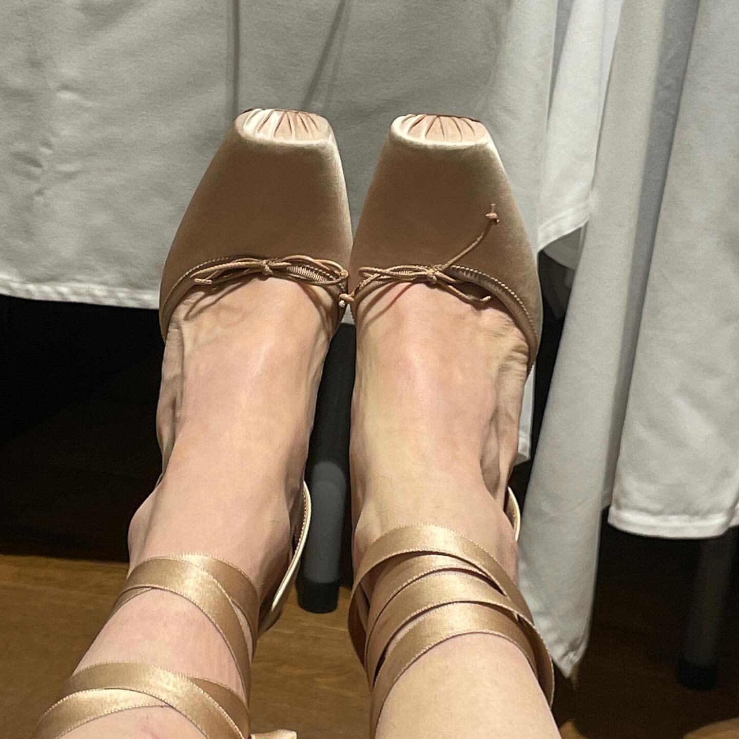 Close-up view of adult female feet wearing light brown ballet-style heels with ribbons, against a neutral background with soft lighting.