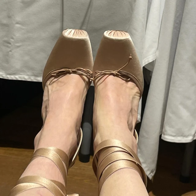 Close-up view of adult female feet wearing light brown ballet-style heels with ribbons, against a neutral background with soft lighting.