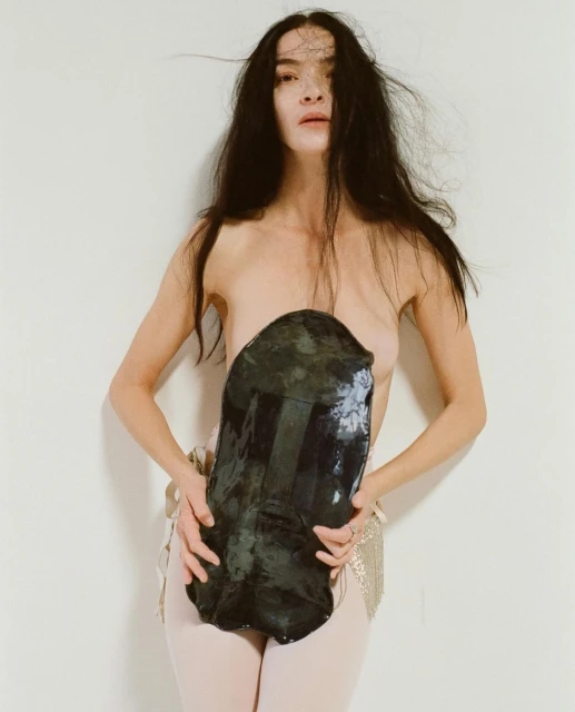 Portrait of a bare-chested adult woman with long dark hair holding a large, dark stone in front of her against a white backdrop.