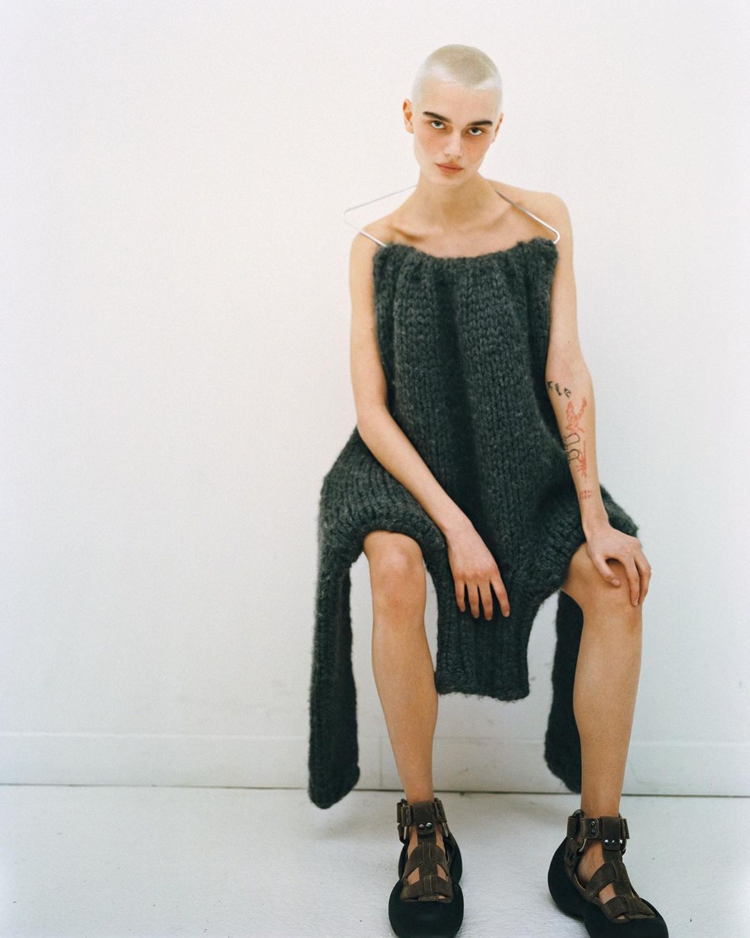 A bald woman in a chunky knit dress and platform sandals sits in front of a white wall. The shot is brightly lit and minimalist.