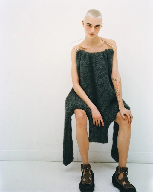 A bald woman in a chunky knit dress and platform sandals sits in front of a white wall. The shot is brightly lit and minimalist.