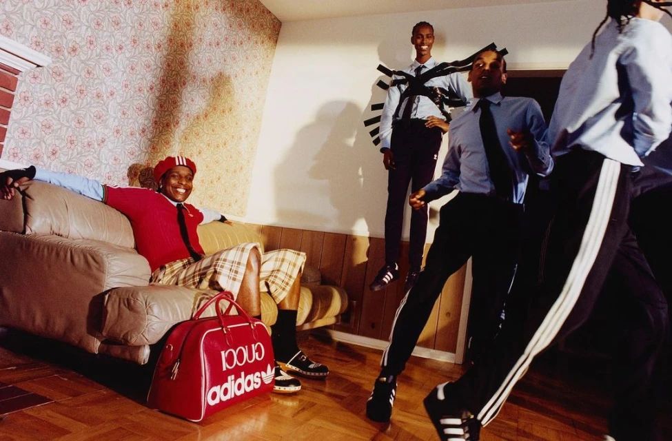 Men wearing Adidas x Gucci apparel in a decorated room. One man reclines on a beige couch near a red Adidas x Gucci duffel bag.