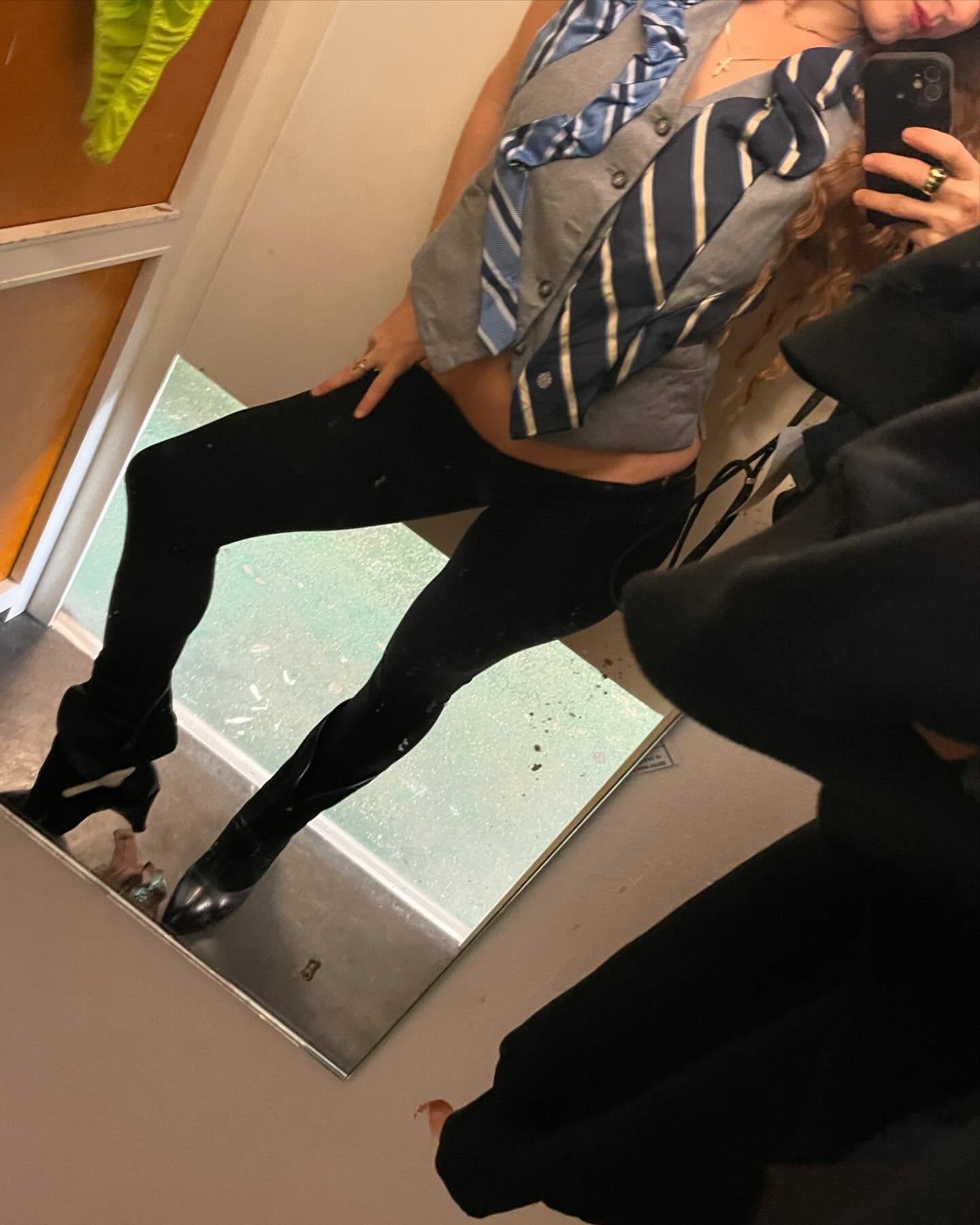 Woman posing in a mirror wearing a cardigan decorated with neckties, black leggings, and heeled boots in an indoor setting.