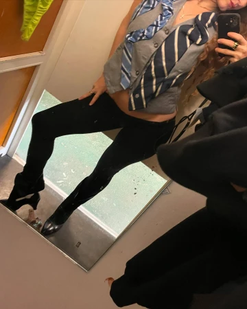 Woman posing in a mirror wearing a cardigan decorated with neckties, black leggings, and heeled boots in an indoor setting.