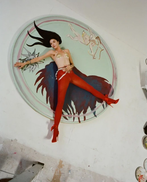 Woman in seashell bra, red tights, and heels poses against a round wall art piece in an indoor setting with a tilted camera angle.