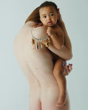 An adult holding a baby who is wearing a charm necklace, photographed against a plain light background by Brianna Capozzi.