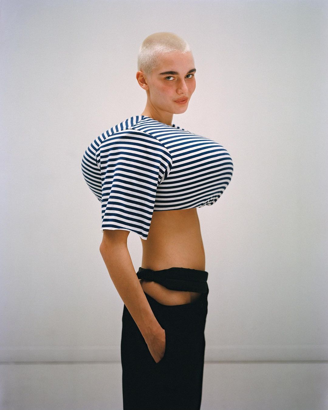 Woman with shaved head poses in a striped crop top and black pants against a white backdrop in a fashion portrait image.