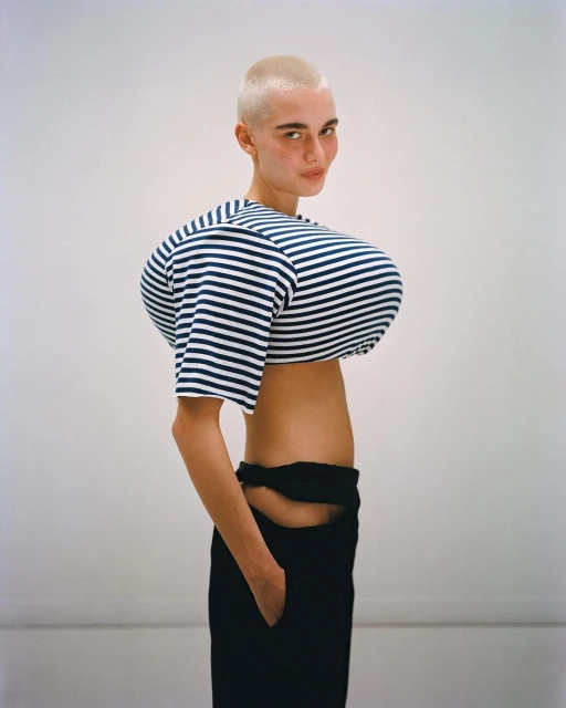 Woman with shaved head poses in a striped crop top and black pants against a white backdrop in a fashion portrait image.