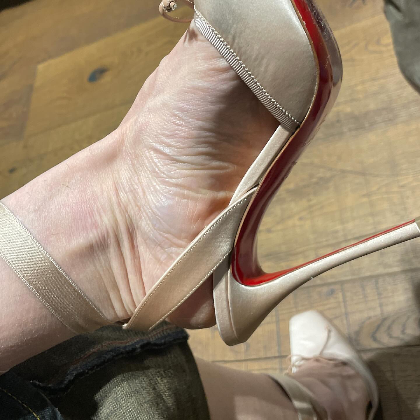 Closeup of a woman's foot in a champagne-colored, high-heeled sandal with a red sole; visible indoors with a wood floor.