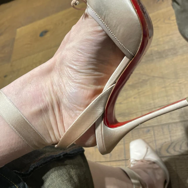 Closeup of a woman's foot in a champagne-colored, high-heeled sandal with a red sole; visible indoors with a wood floor.