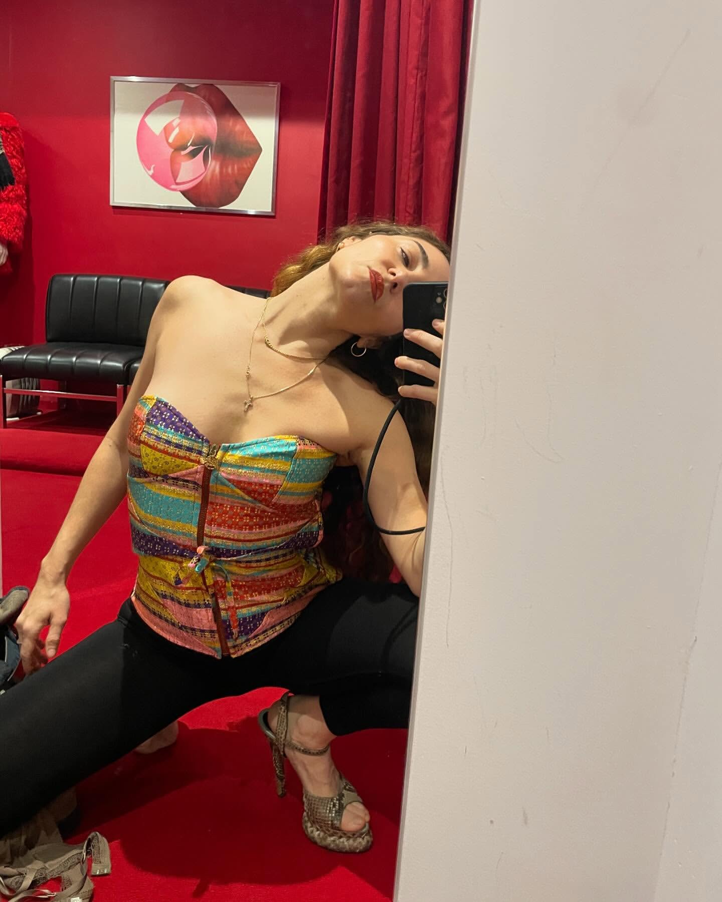 Woman in patterned top and black leggings poses in front of a mirror, with red wall and black chair in the background. Indoor setting.