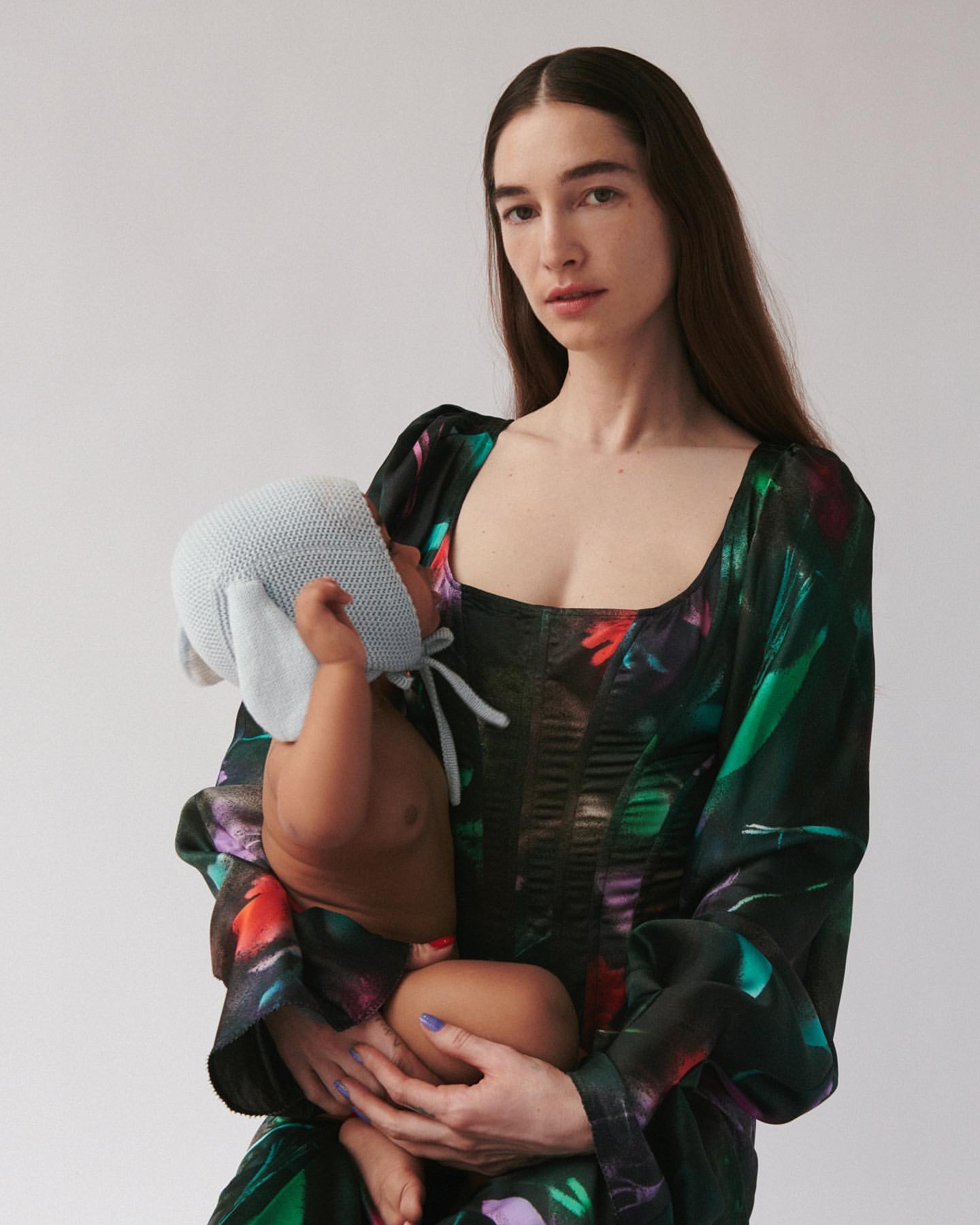 A mother with long dark hair holds her baby, who is wearing a light blue bonnet, against a light background. The mother wears a floral dress.
