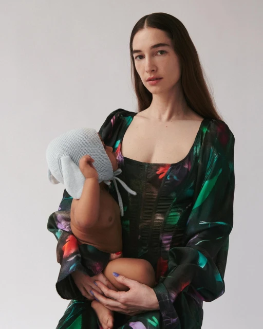 A mother with long dark hair holds her baby, who is wearing a light blue bonnet, against a light background. The mother wears a floral dress.