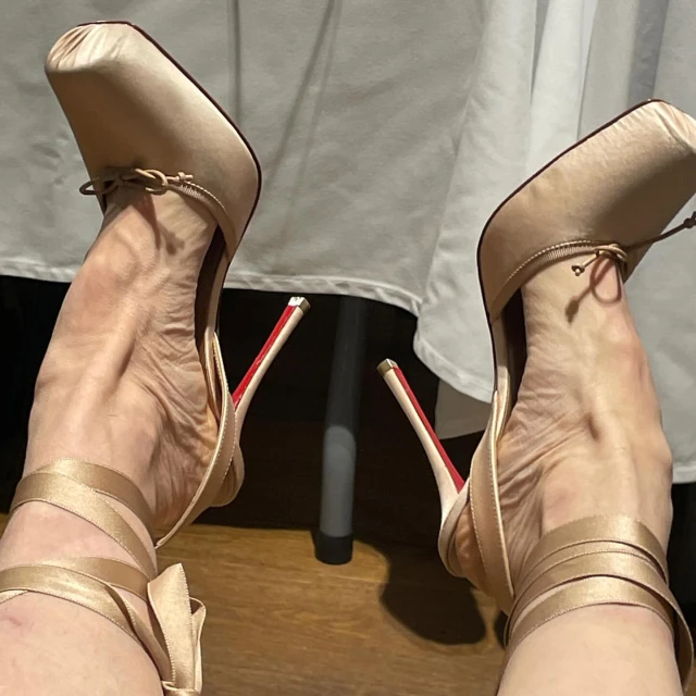 Close-up of a person's feet wearing tan-colored high heels with ribbon ankle straps and red soles, set against a neutral background.