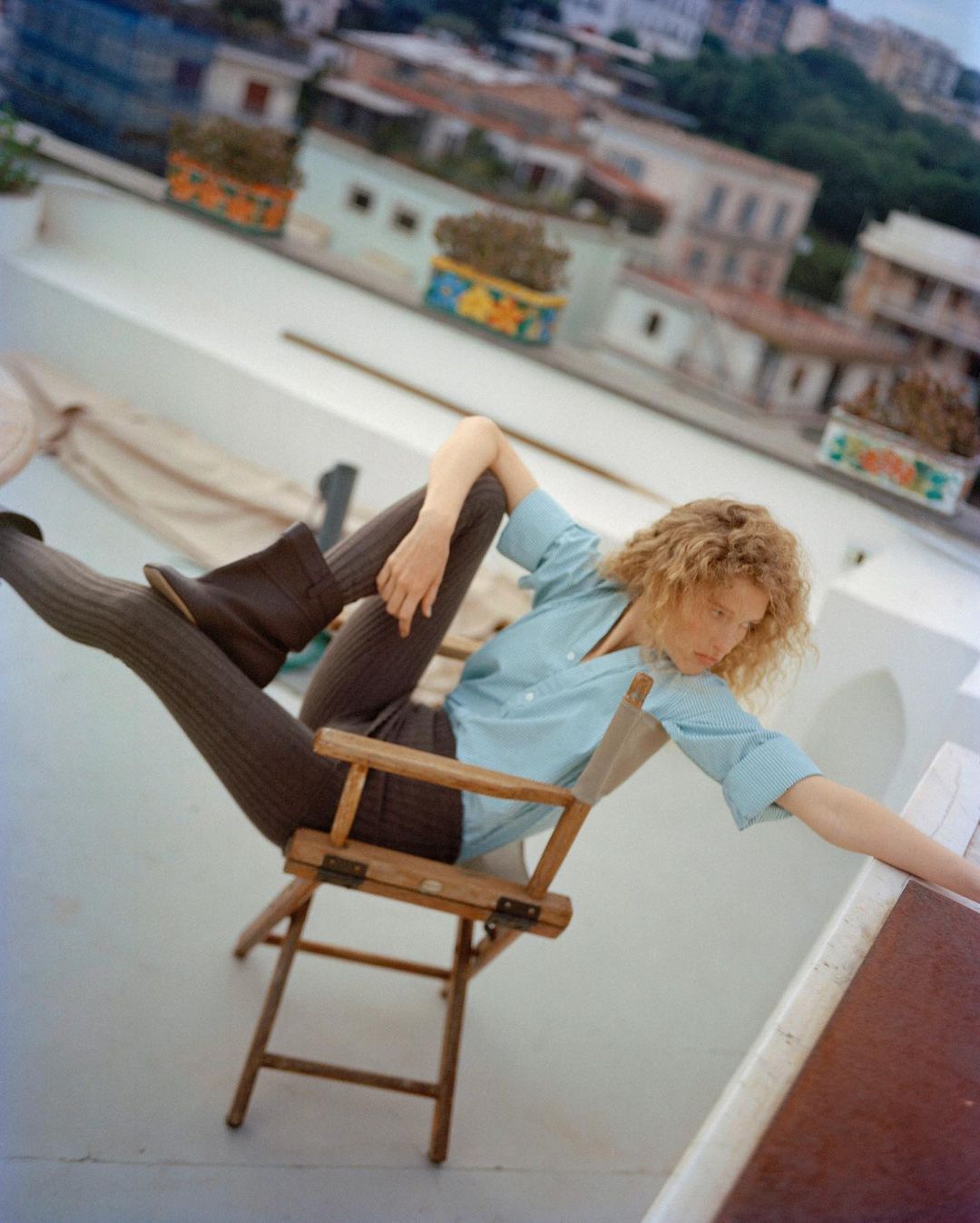 Woman in a light blue shirt and grey leggings sitting backward on a wooden chair on a rooftop with a cityscape background.