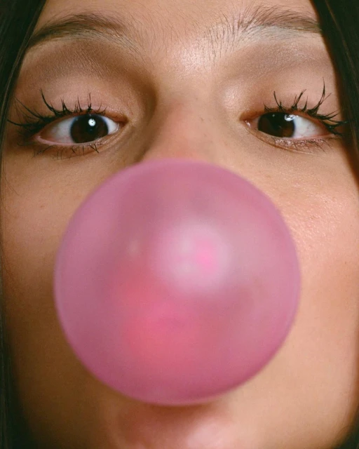 Extreme close-up of a woman with long eyelashes blowing a large pink bubblegum bubble, eyes and face filling the entire frame.
