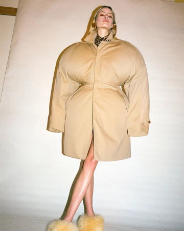 Model wearing an oversized tan puffer coat and furry yellow slippers against a plain cream studio background.