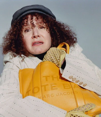Woman with curly hair in newsboy cap holds a yellow Marc Jacobs tote bag in a studio portrait against a neutral background.