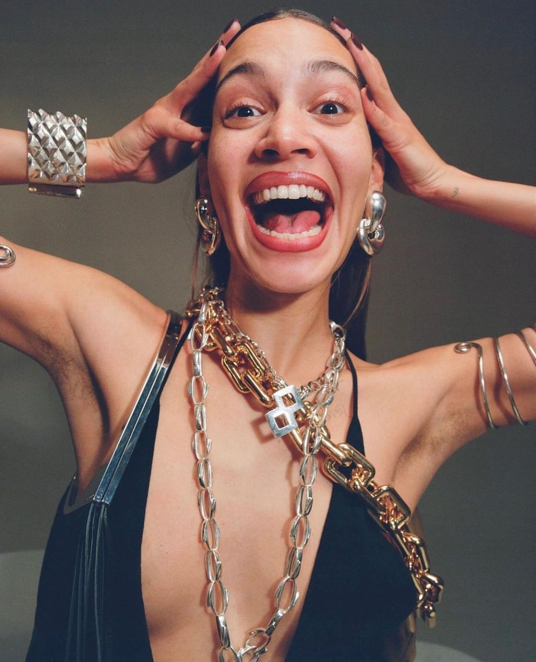Woman with an open mouth, wearing multiple metal necklaces, a black top, and holding her hands on her head in an indoor setting.