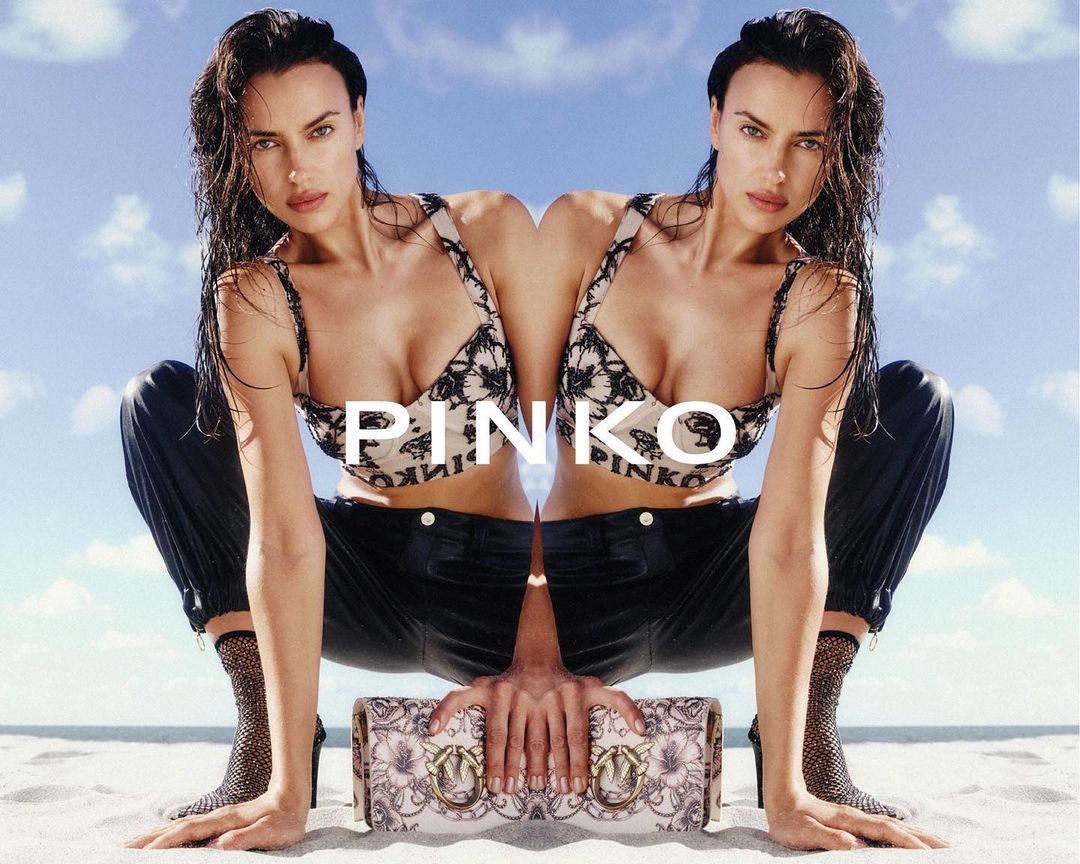 Mirrored image of a woman in a bralette and pants, holding a purse, on a sandy beach against a bright sky with few clouds.