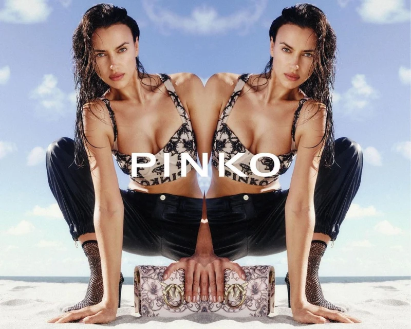 Mirrored image of a woman in a bralette and pants, holding a purse, on a sandy beach against a bright sky with few clouds.