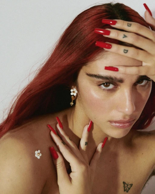 Close-up portrait of an adult woman with red hair, red nails, and a butterfly tattoo, against a neutral background. Fashion shot.