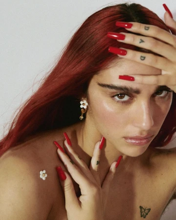 Close-up portrait of an adult woman with red hair, red nails, and a butterfly tattoo, against a neutral background. Fashion shot.