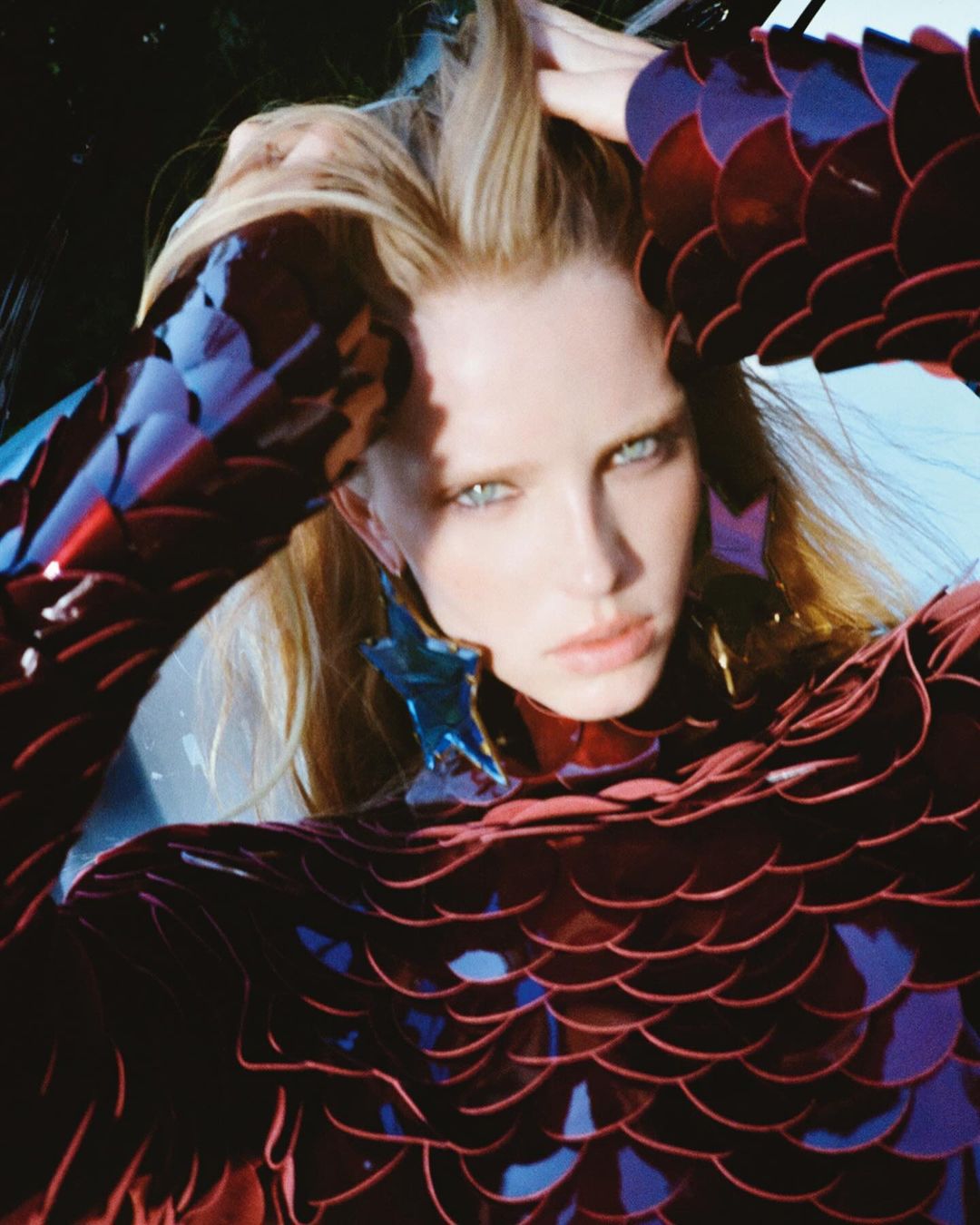 Woman with blonde hair poses in a dark-red scale-patterned dress with star-shaped earrings against a blue sky background in a medium shot.