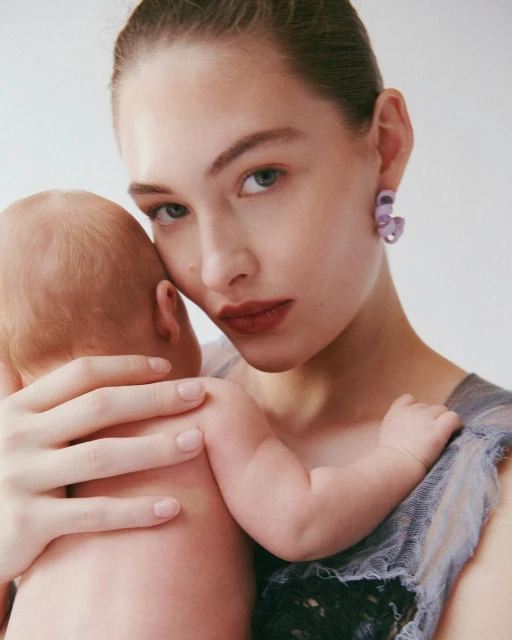 Close-up of fair-skinned young woman holding baby against a plain backdrop. Woman wears earrings and sheer, draped garment; baby is swaddled.