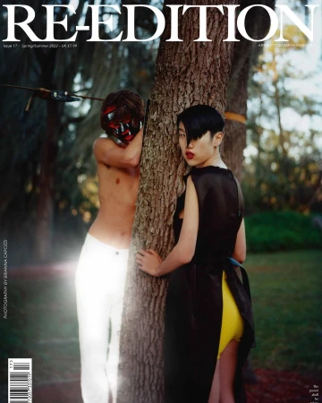 Magazine cover featuring two models: one partially obscured by a mask, and one beside a tree in a sheer black top and yellow shorts.