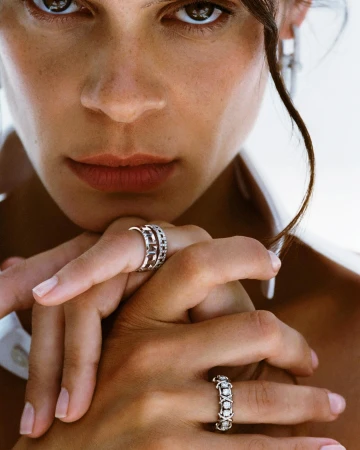 Close-up of an adult female with light skin and brown hair modeling two silver rings in a bright indoor fashion setting.