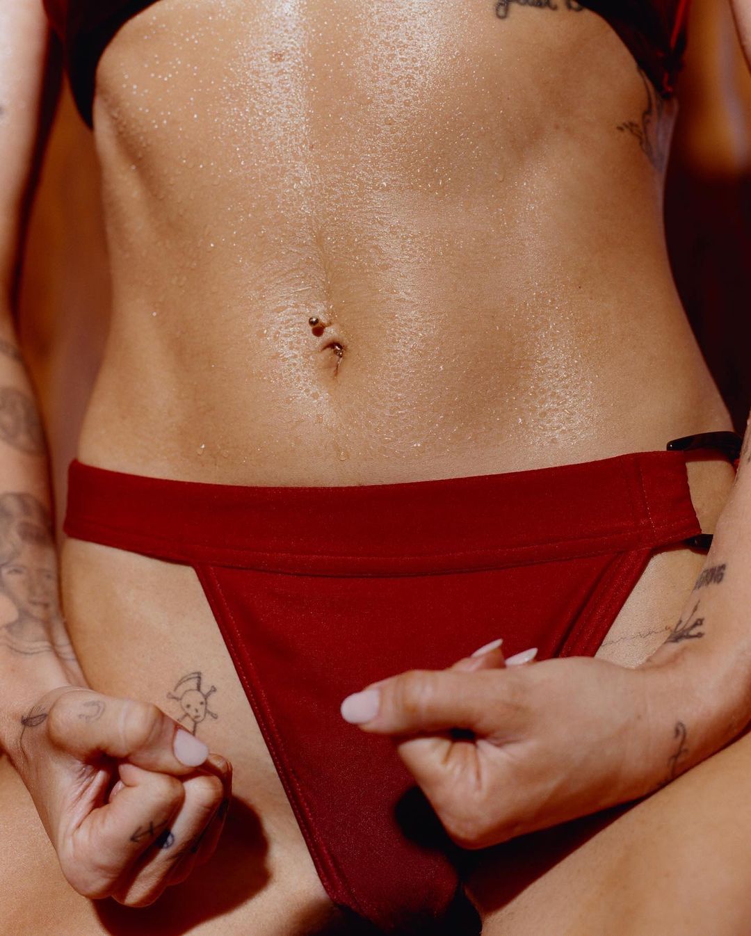 Close-up of a woman's torso with a slim figure, visible navel piercing, and tattoos. She wears a red bikini bottom and bra.