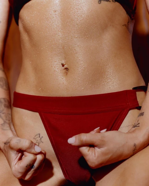 Close-up of a woman's torso with a slim figure, visible navel piercing, and tattoos. She wears a red bikini bottom and bra.