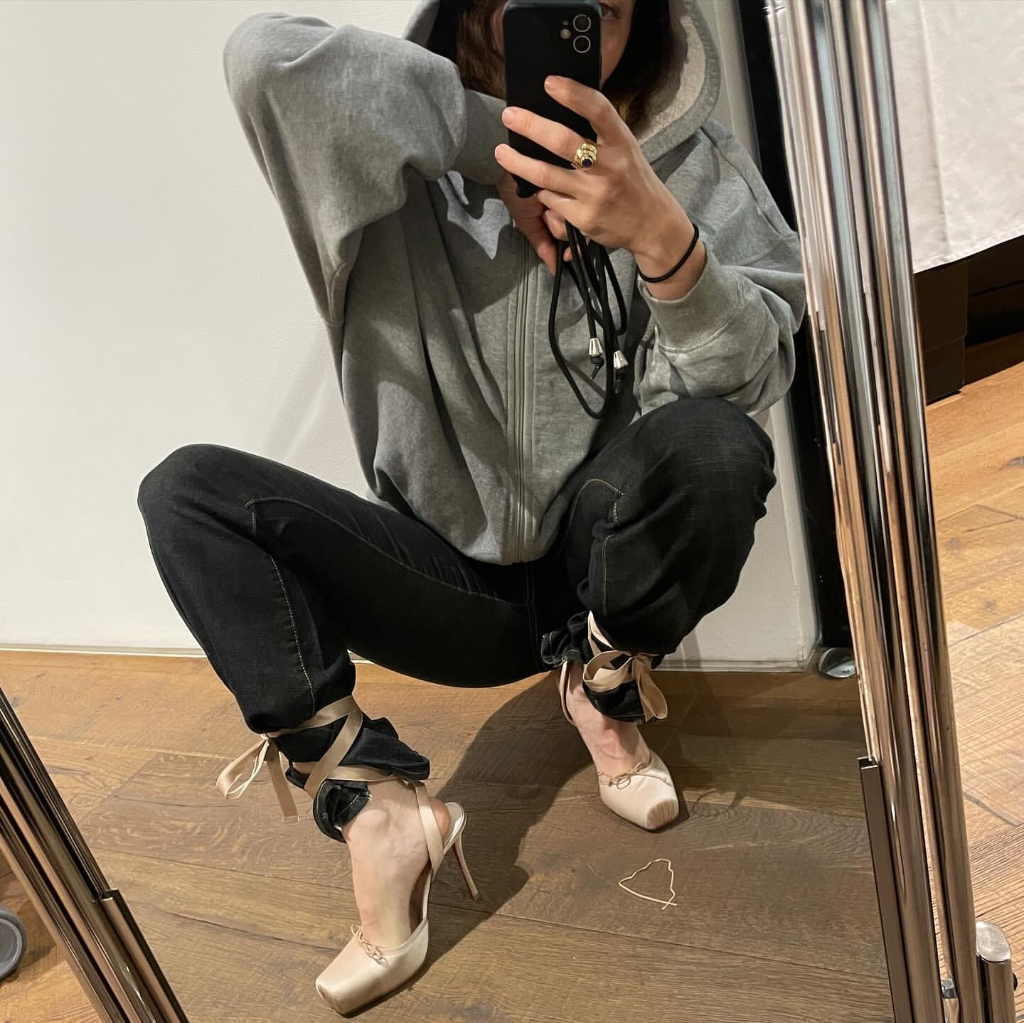 Young woman takes a mirror selfie while squatting, wearing a gray hooded sweatshirt, black jeans, and beige high heel shoes with ribbons.