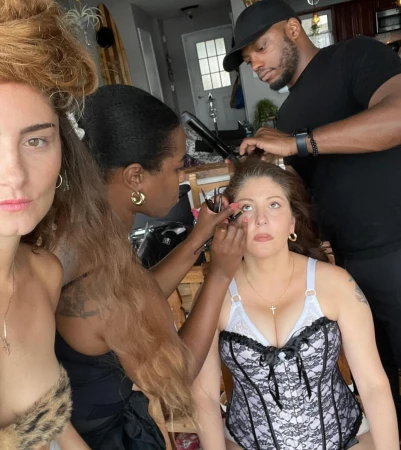 Behind-the-scenes shot of a makeup artist applying makeup to a model while another stylist arranges her hair, inside an airy room.