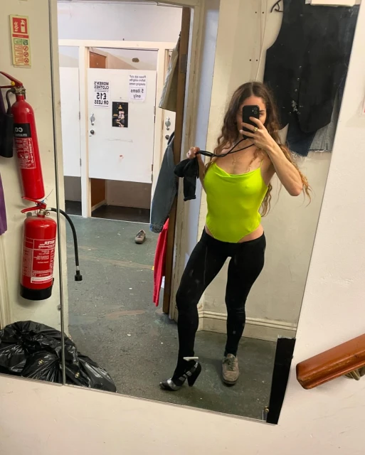 Woman in a neon green top and black leggings takes a mirror selfie in a grungy indoor setting with a fire extinguisher visible.