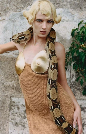 Fashion shot of a blonde woman wearing a gold dress with a boa constrictor snake around her neck, posing against a stone wall.