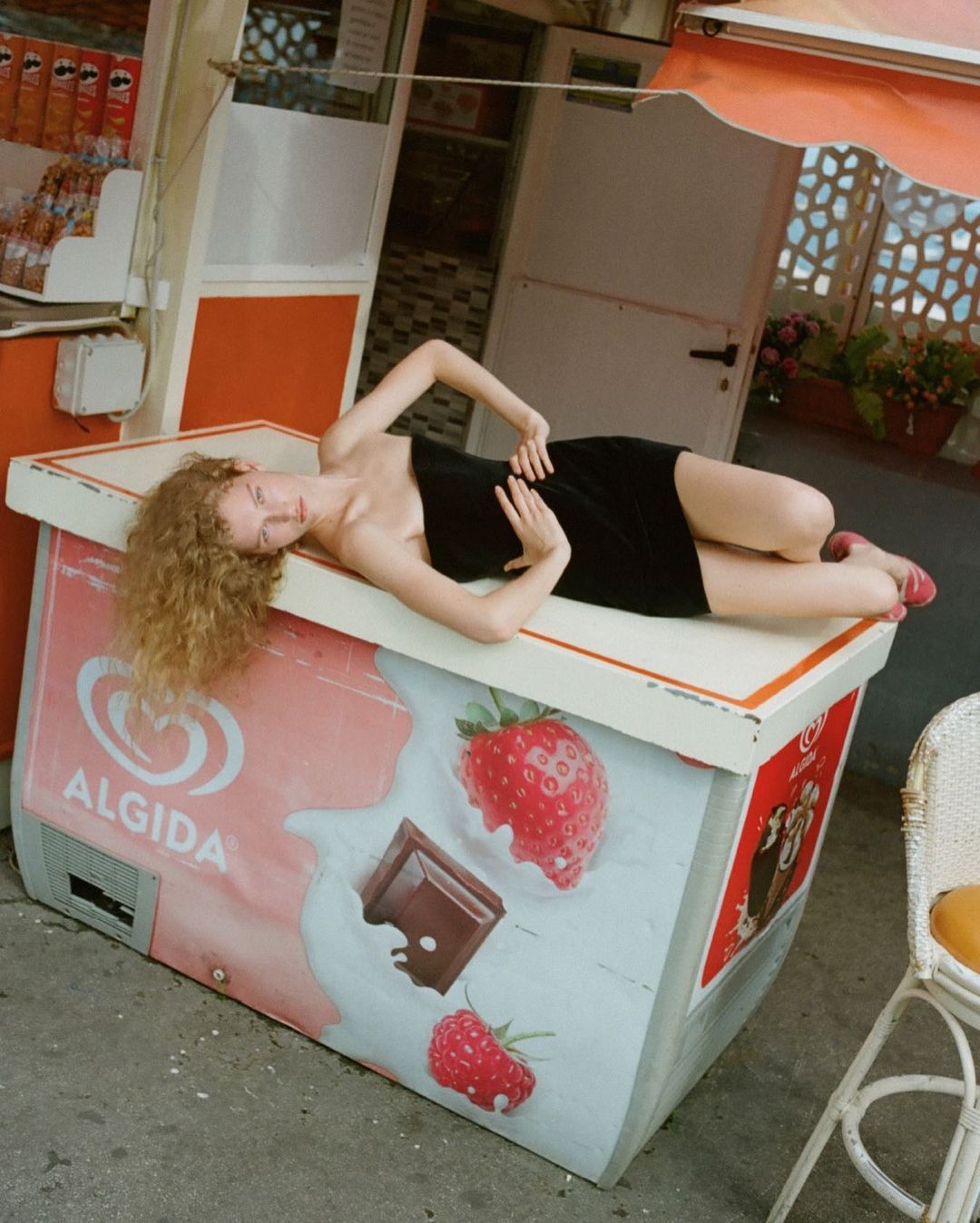 A woman in a black dress reclines on a strawberry-decorated ice cream freezer in front of a storefront with sunlight streaming in.