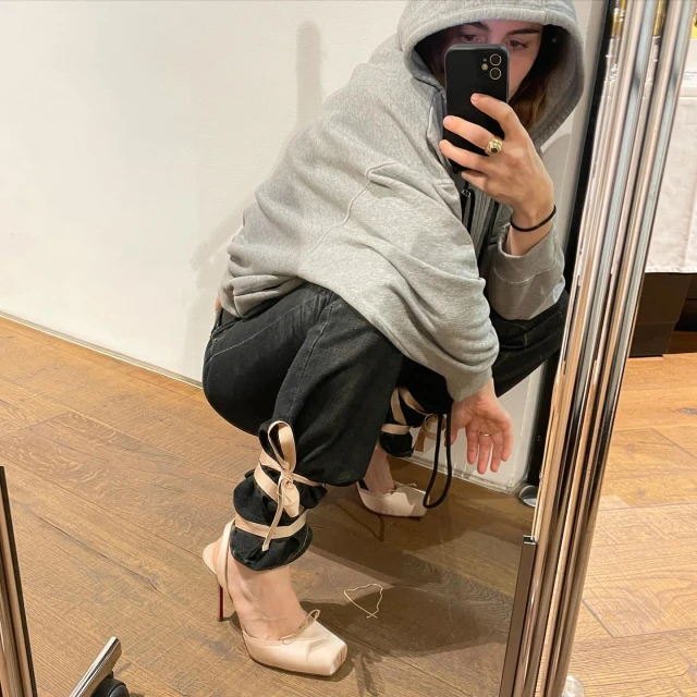 Woman in a grey hoodie and black jeans poses while taking a mirror selfie, showcasing ivory ballet-style high heels with ribbons.