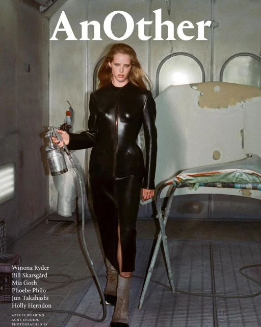 Fashion editorial image featuring a model in a black leather outfit holding a spray gun in an auto paint shop, magazine cover 'AnOther'.