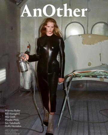 Fashion editorial image featuring a model in a black leather outfit holding a spray gun in an auto paint shop, magazine cover 'AnOther'.