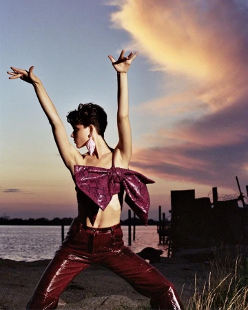 Fashion shot of a young adult woman posing in a purple top and pants with arms raised against a sky with colorful sunset clouds.