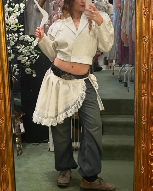 Indoor mirror selfie of an adult female wearing a white cropped jacket, wrap skirt, denim pants, and brown shoes, holding a clothes hanger.