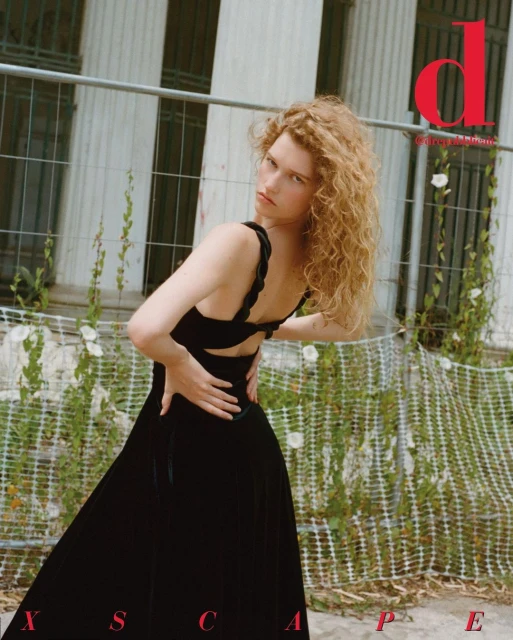 Fashion shot of a young adult woman with curly blonde hair wearing a black dress. She is posed in front of a building.